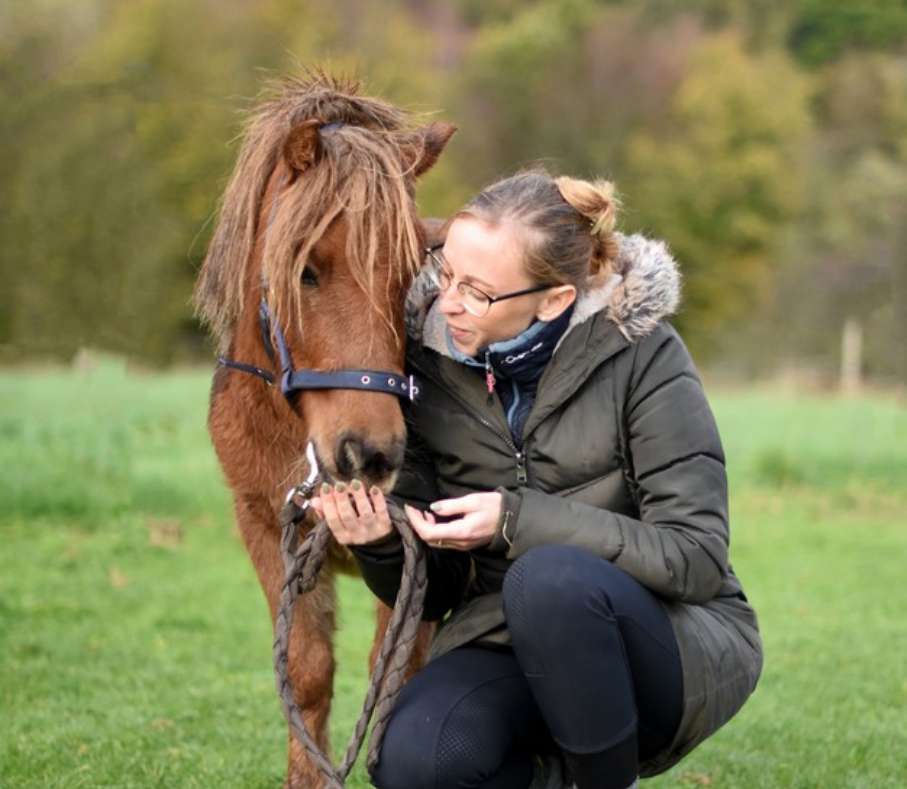 Amélie Focant est accroupie à côté d'un poney brun et elle lui fait sentir sa main