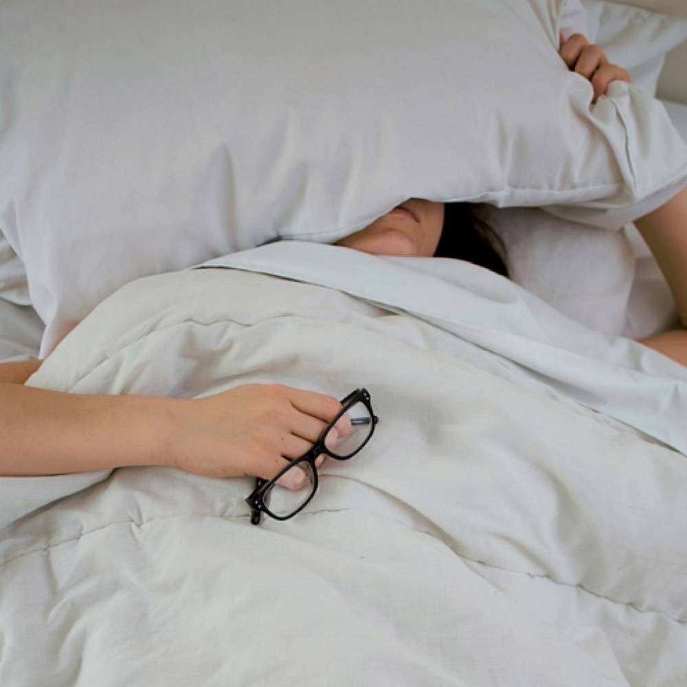 Une personne allongée sous une couette blanche avec une main qui tient le coussin et l'autre qui tient des lunettes