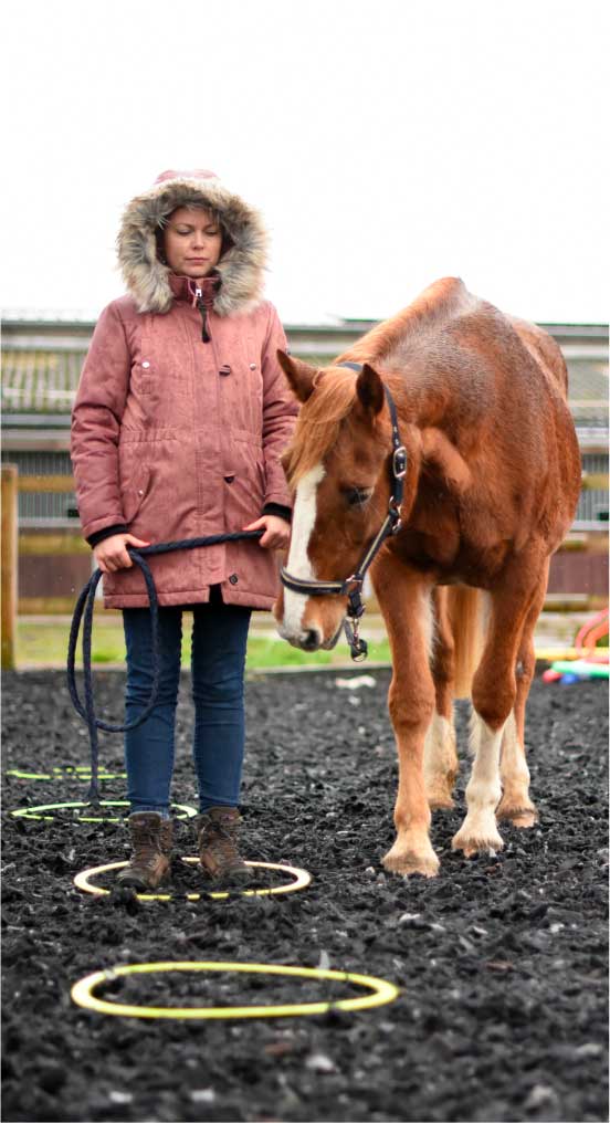 Une femme portant un manteau rose et une capuche à fourrure se tient debout à côté d'un cheval alezan