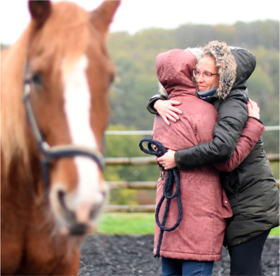 Deux femmes se donnent une étreinte dans un manège avec un cheval à la robe alzane et sa crinière blanche flou en premier plan lors d'une formation chez EquiLink