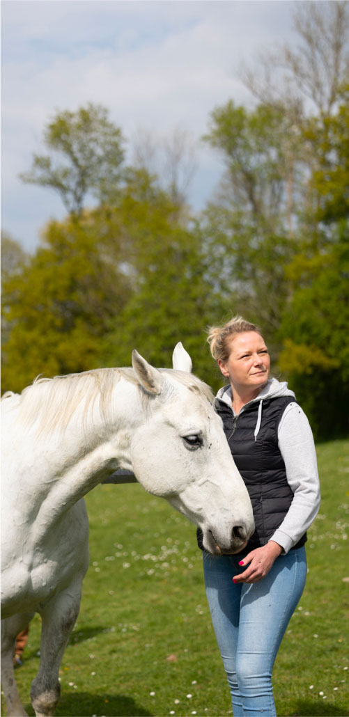 Une dame aux cheveux blonds se tient droite à côté d'un cheval blanc